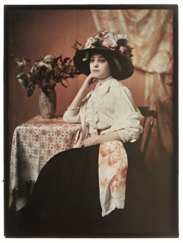 Autokrom, portrett av en sittende kvinne ved et bord. Hun bærer en stor hatt med blomster og det er blomster i vasen ved siden av.